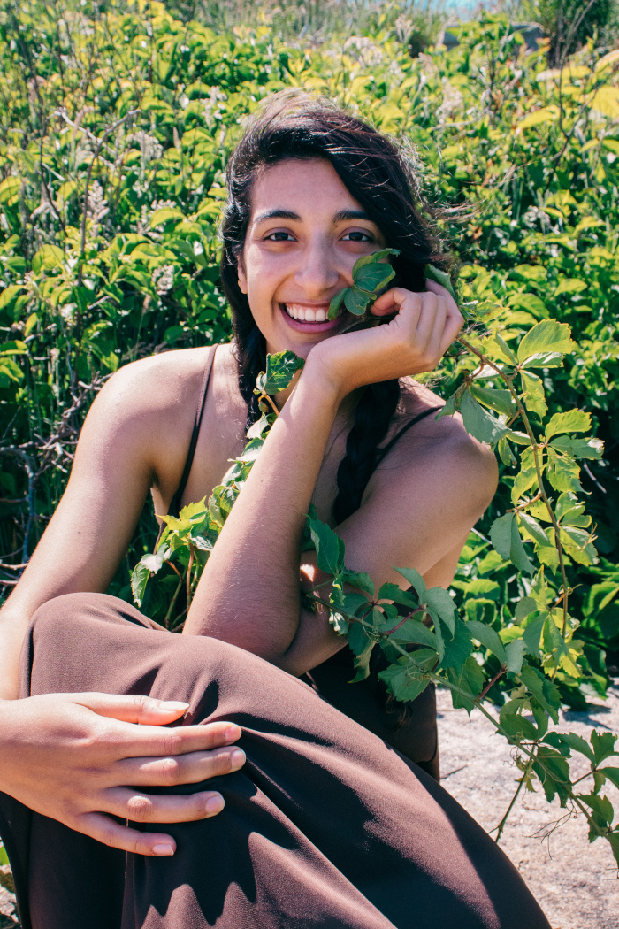 Cassandra Myers in a brown dress and smiling widely, surrounded by greenery and holding a branch with leaves near their face