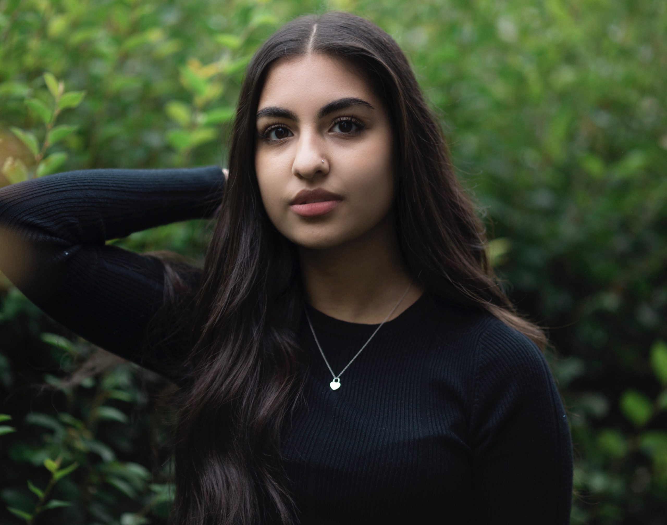 an image of Alysha Mohamed in a ribbed black shirt and a small silver necklace, standing in front of lush greenery with one arm up and bent.