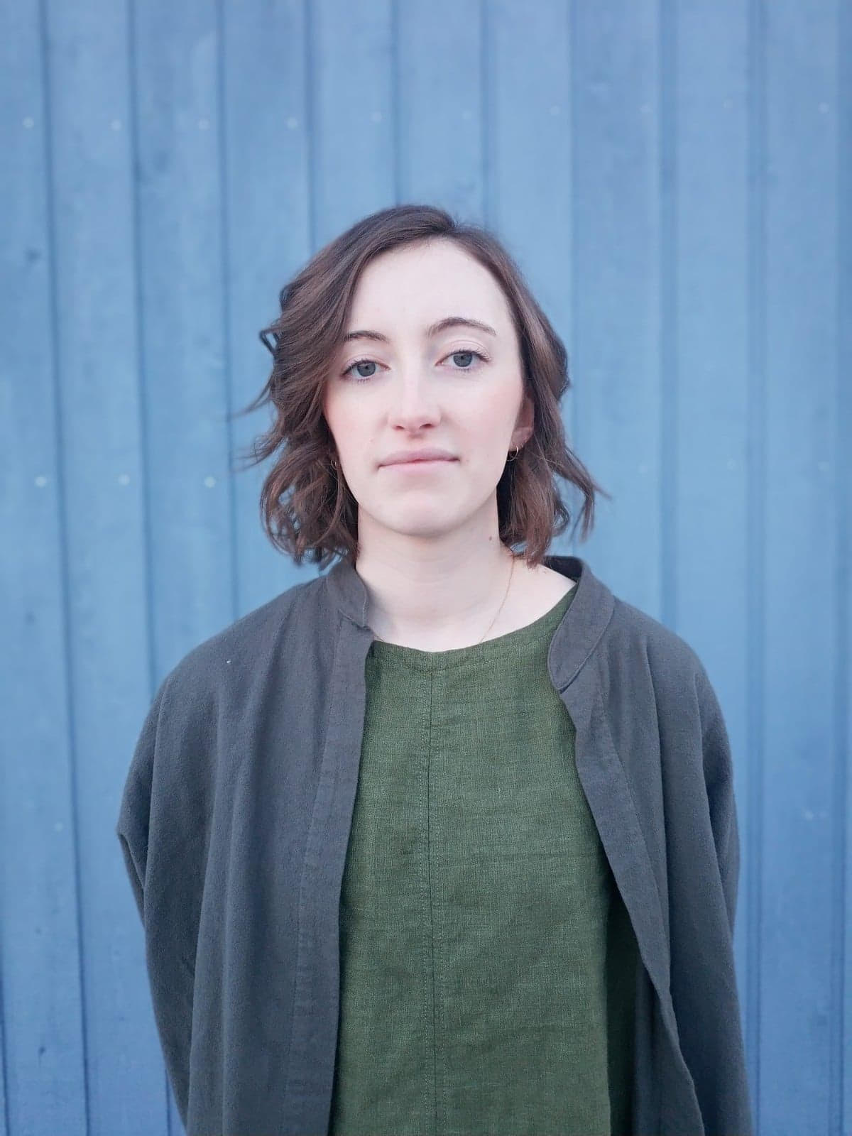an image of Alison Braid standing in front of a blue wall in a simple green shirt and black overshirt.