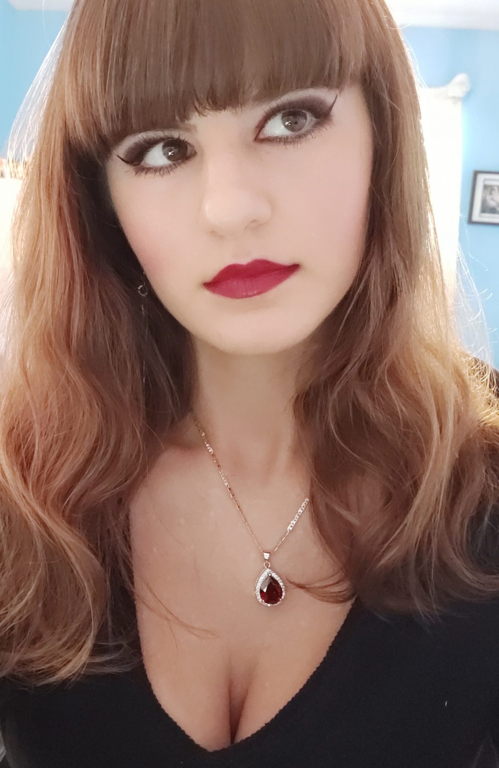 Noelle Brody has long red-brown hair with bold bangs that end just above her eyes, as well as red lipstick, thick eyeliner, a necklace with a red stone and a black top. Her eyes looks off to the right but se is facing the camera directly.