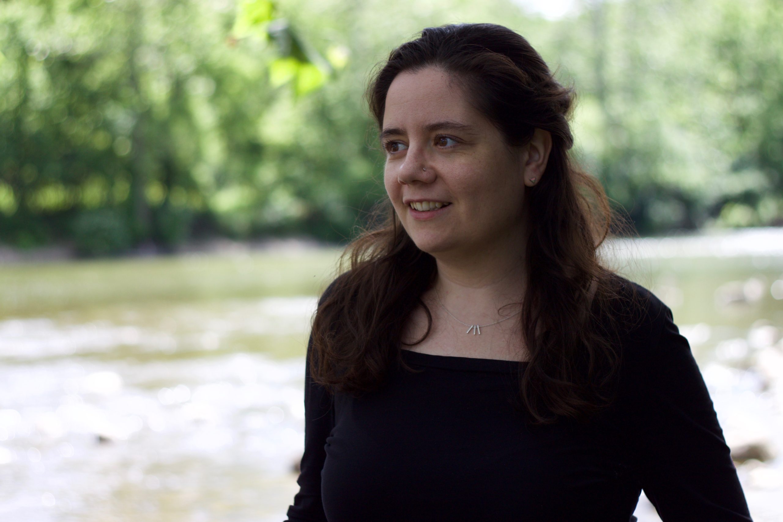 an image of Annick MacAskill in a black shirt and sweater with a small silver necklace, standing in front of a river and looking off to the side