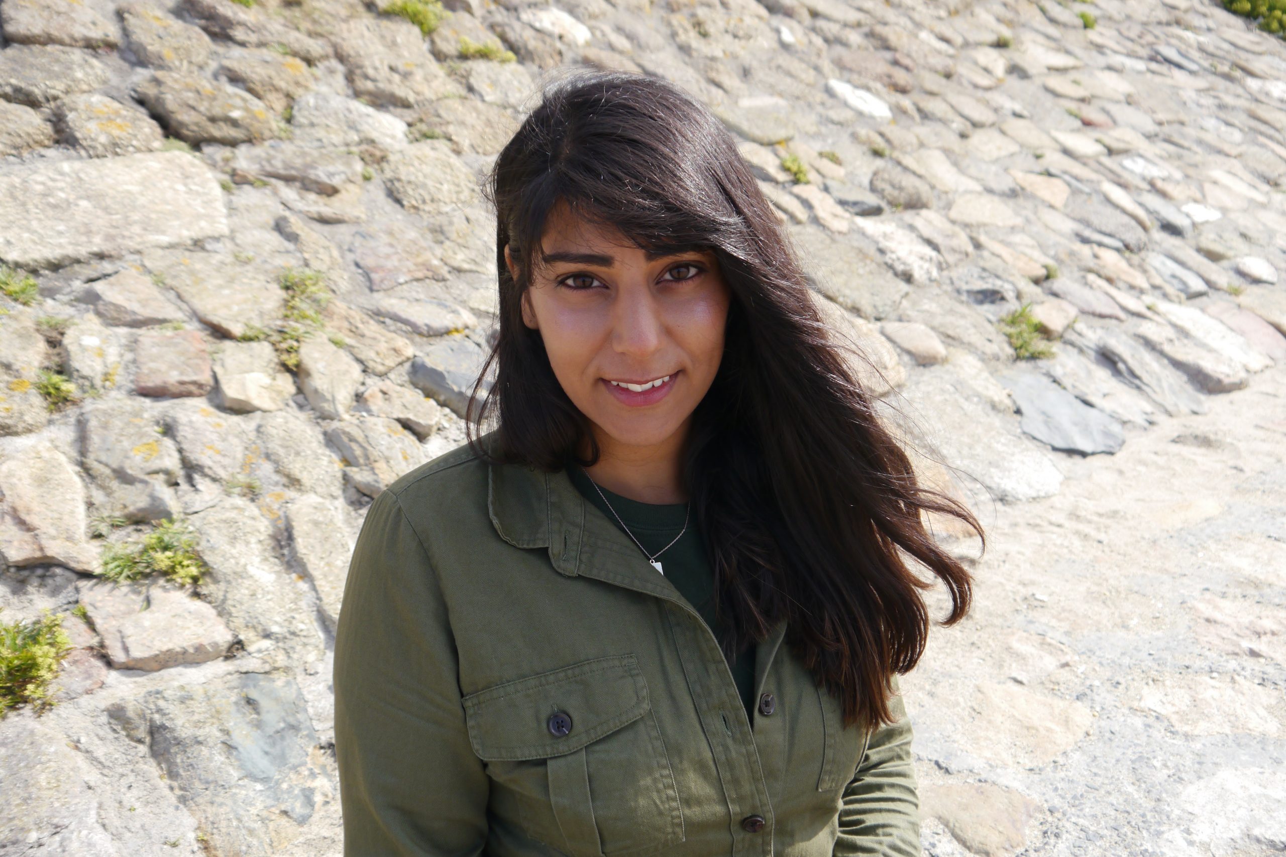 an image of Alycia Pirmohamed in a light coat, hair down, standing in front of a cobblestone wall.