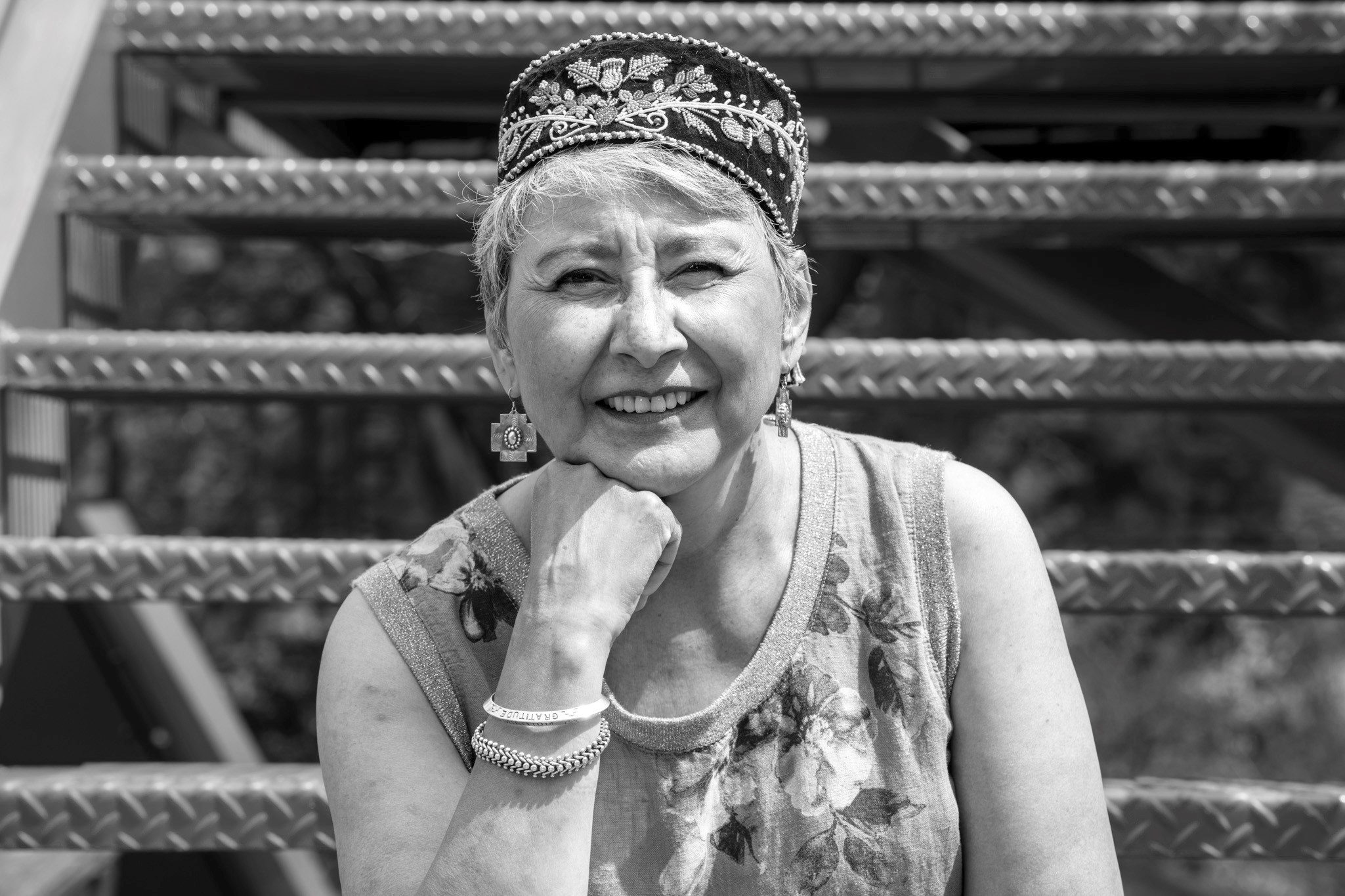 a black and white image of a person with white hair in a tank top with flowers printed on it, two bracelets, large square-ish earrings, and a circular hat with embroidered leaves and flowers; she is sitting on an outside staircase, smiling and squinting with one hand supporting her chin.