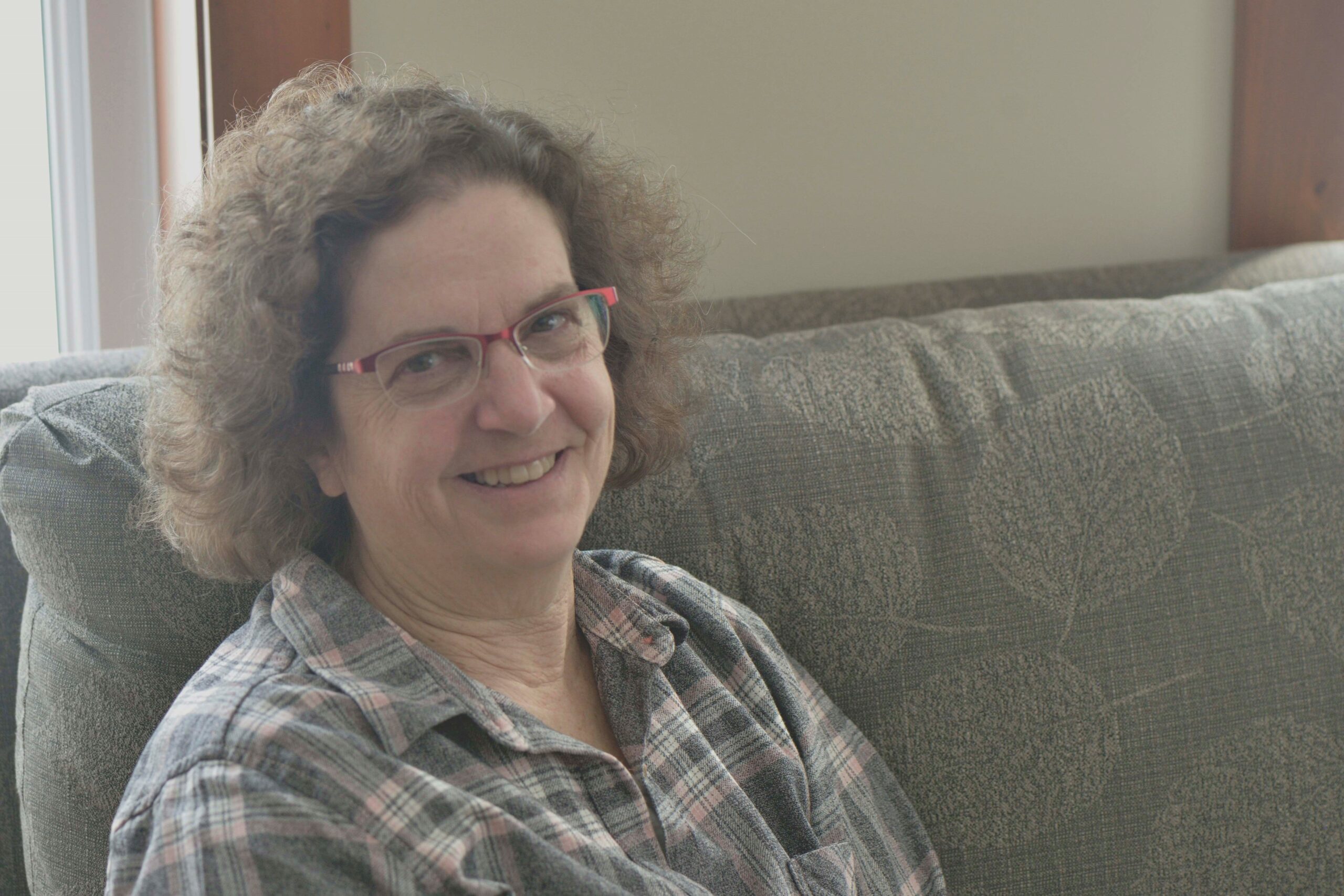 Anne Marie Todkill is wearing a plaid shirt and red-framed glasses, with curly hair and a smile.