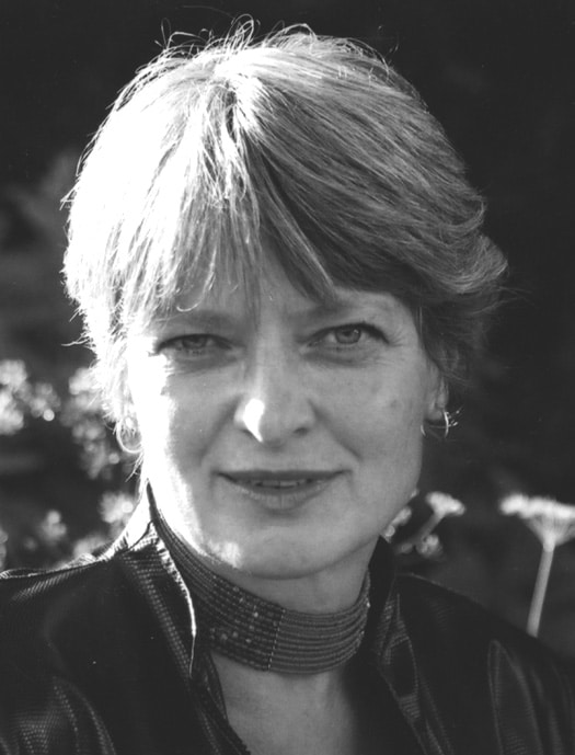 a black and white photo of Di Brandt's face in beaded chocker necklaces and a dark jacket, with short hair and a direct gaze