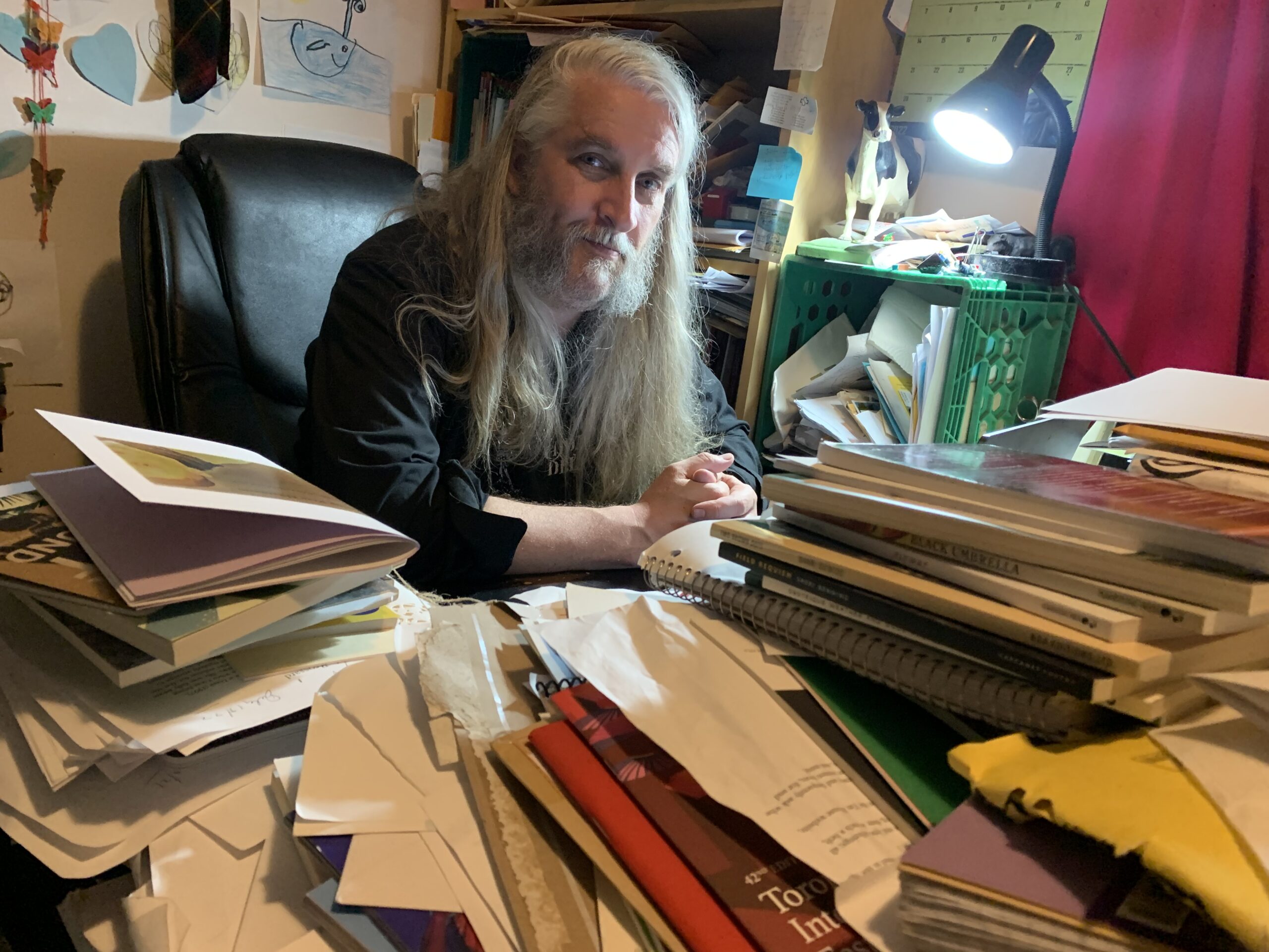 rob mclennan, with long hair grey hair, a short bushy beard, and a serious expression has been photographed across a mound of books and papers and chapbooks and zines; the photo is also at a slight dutch angle, which makes the perspective/the subject feel off-kilter and odd