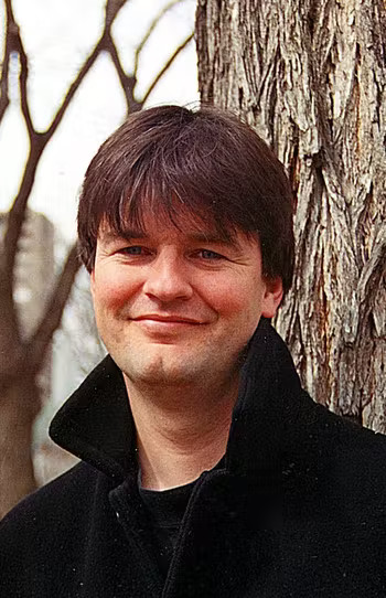 Tim Bowling stands in front of a tree, beside a larger tree trunk, in a black coat. He is smiling and has brown hair with bangs.