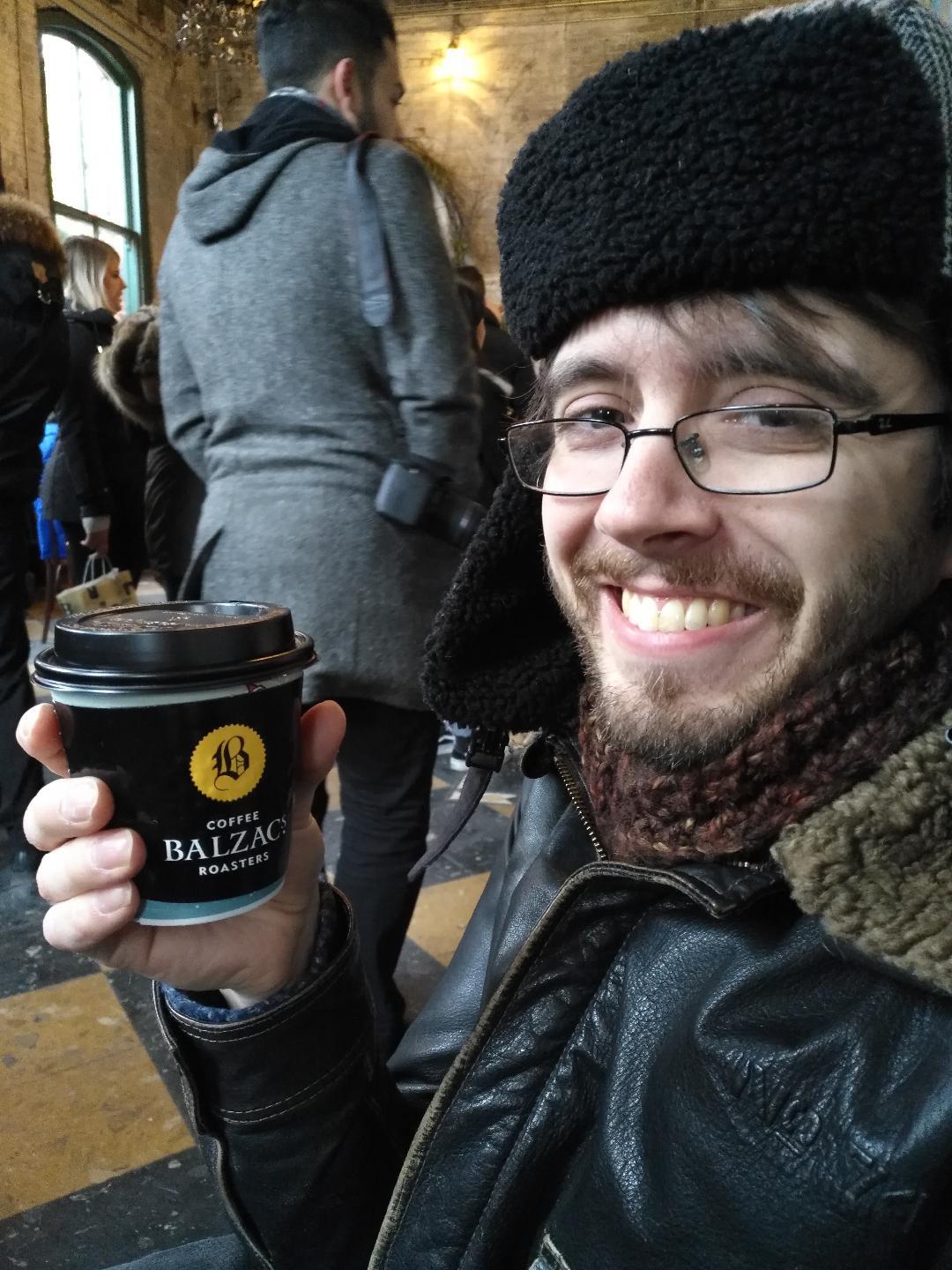 David Barrick wears thin-rimmed black glasses and a furry hat, with a sparse beard and mustache and a paper coffee cup in his hand; he is grinning and is sitting, with people's backs in the background behind him.
