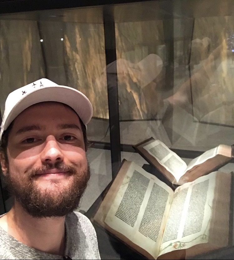 Joseph Kidney stands in front of a display case with an open book inside; he is wearing a white baseball cap and shirt, and has a brown beard and brown hair.