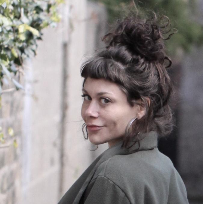 Lianne O’Hara looks back over her shoulder, wearing a dark coat and her brown hair up in a top-of-the-head bun; she smirks, standing beside a brick wall with vines growing on it.