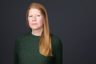 Standing in front of a black background, Lisa Martin has a serious expression and long, straight red hair that comes down and rests on her chest, which is covered by a textured green sweater that compliments her eyes; she has a neutral expression and though she is facing forward, she does not make eye contact with the camera.