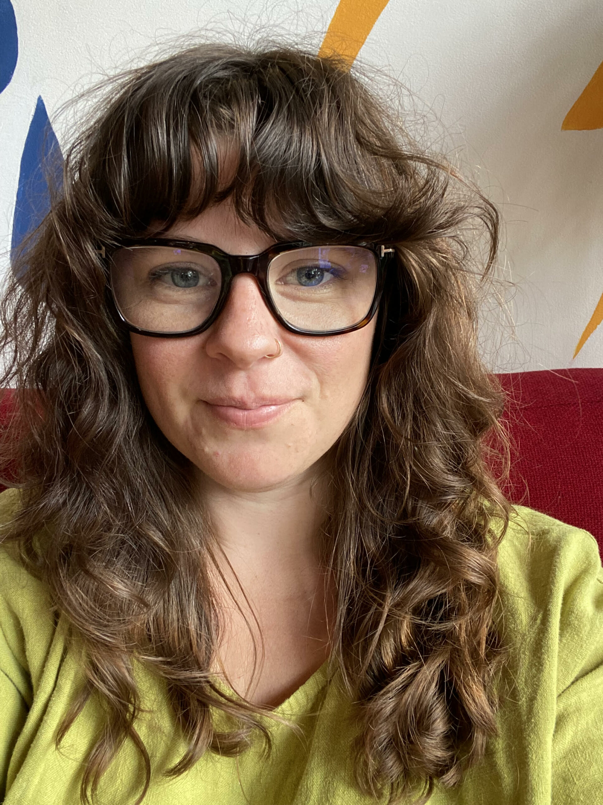Mary Sauer is wearing a yellow top and oval glasses with thick black rims; she sits in front of a white and red background and her hair is long, curly and has bangs.