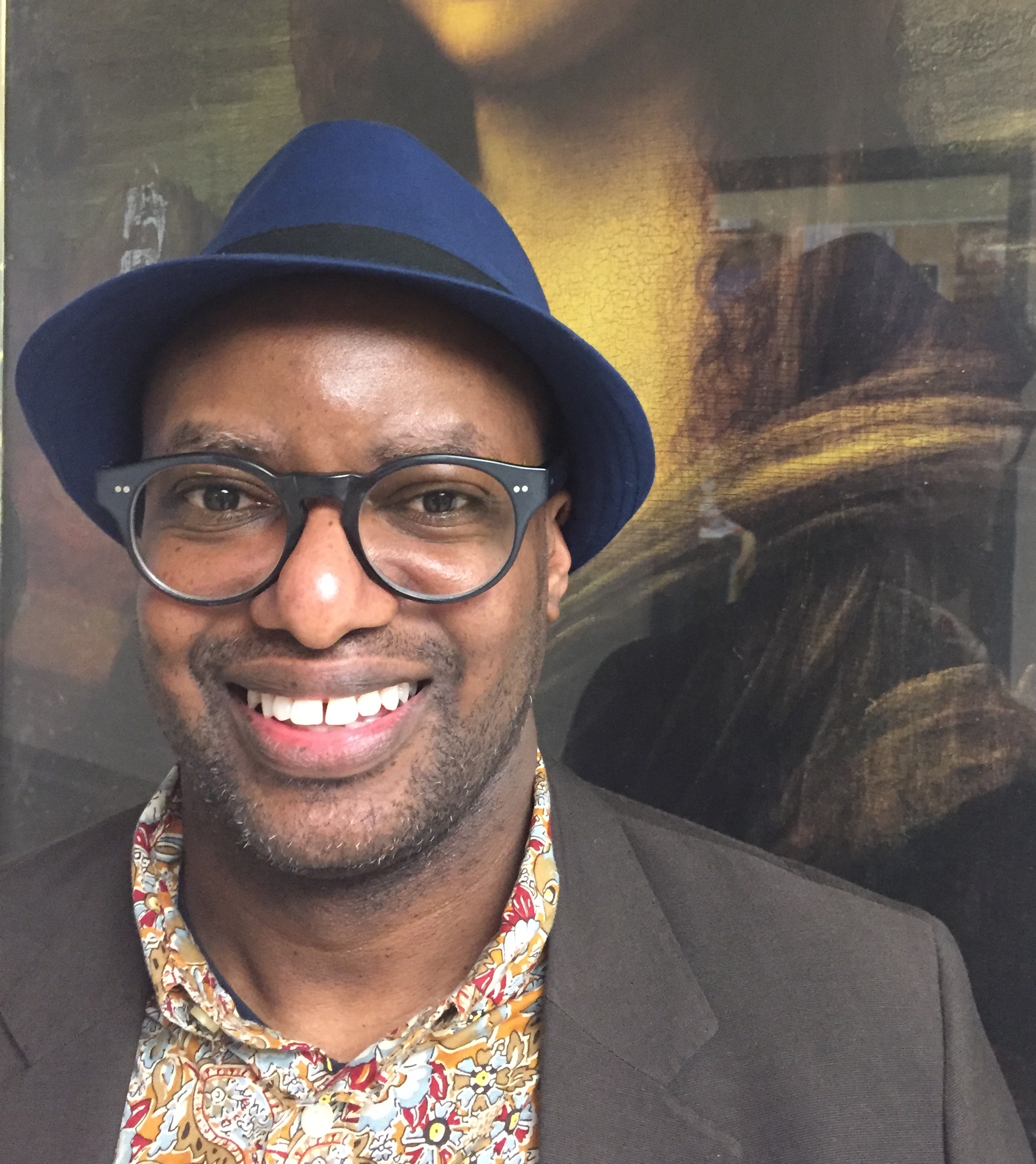 Michael Fraser is seen from mid-chest up, wearing a blue Trilby hat with a dark brown band, a brown suit jacket and a colourfu (mainly orange)l, patterned shirt with a collar. He smiles, looking at the camera in a friendly way through his large, round glasses.
