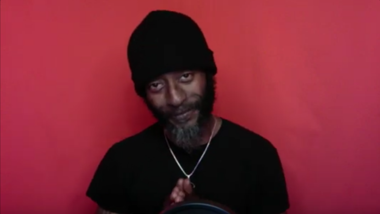 Wakefield Brewster, with his hands together and dressed in black (including a beanie) and a thin gold chain, standing in front of a red background