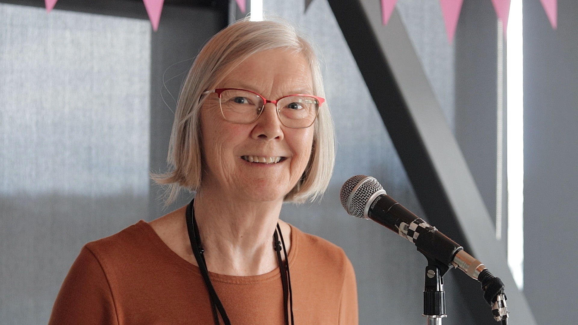 Frances Boyle, in an orange sweater and a long black necklace of many strands, stands in front of the a microphone on a stand; she has chin length white-grey hair and thin-rimmed glasses, and she looks away from the mic to smile at the camera