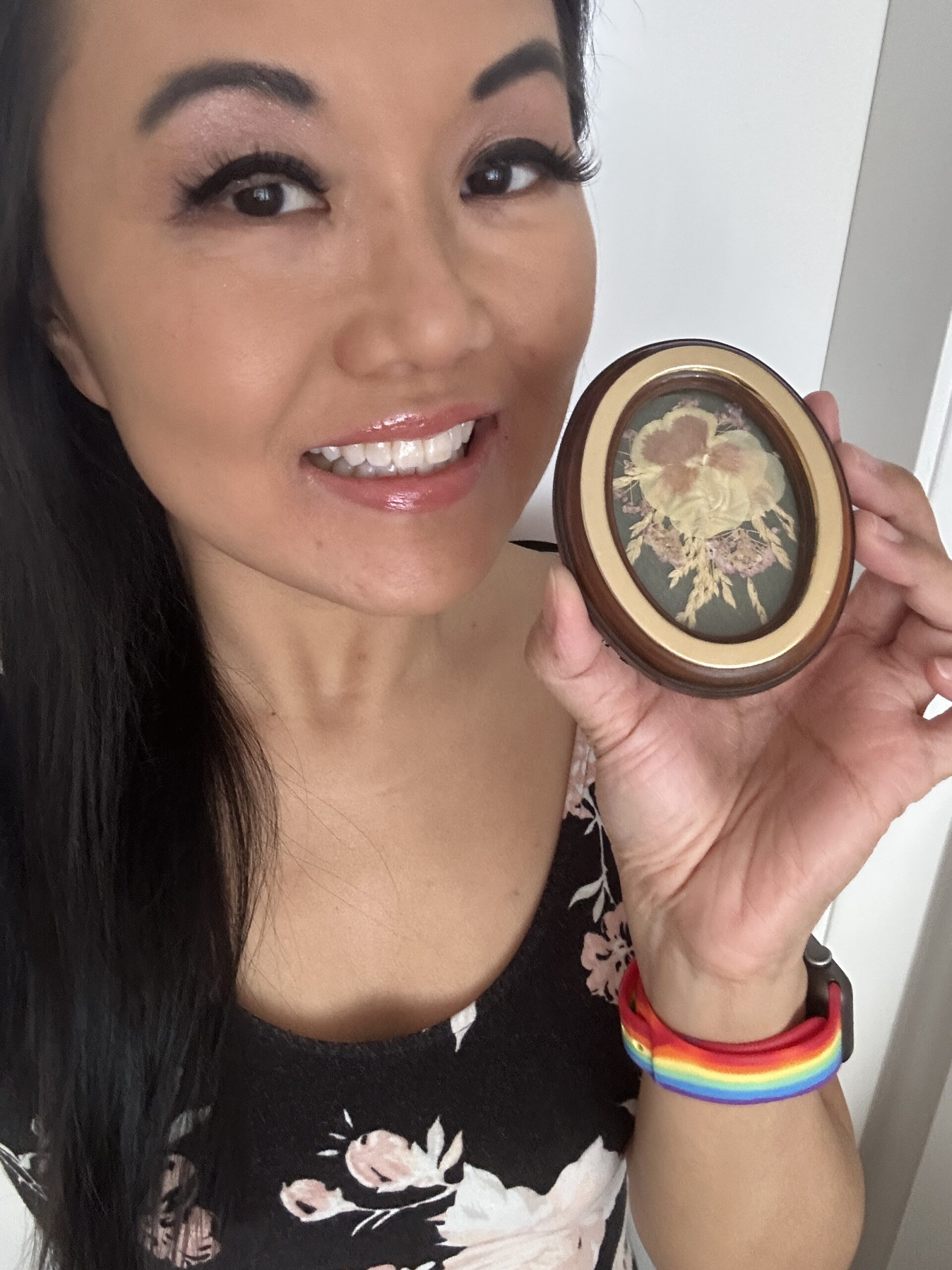 Catherine Lewis has black eye makeup, long black hair, and is holding a small frame with pressed flowers.