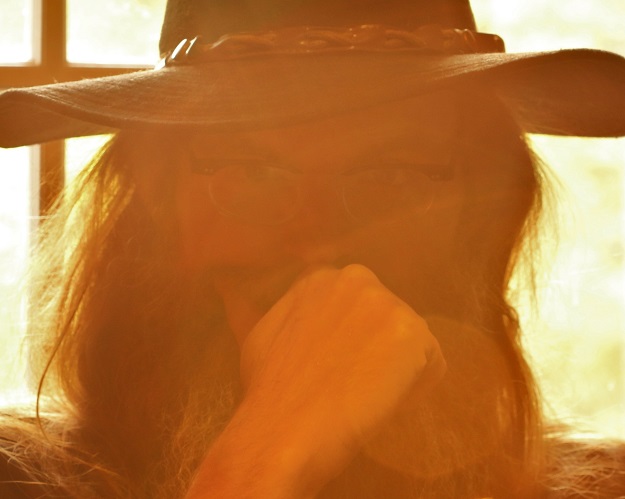 A gold hue image of Matthew King wearing a brimmed hat and partially covering his face with his hand.
