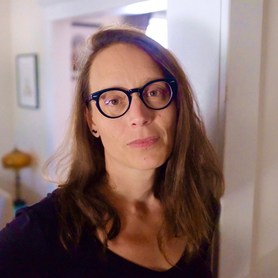 Photo of Brecken Hancock, in a room with light coloured walls. Brecken is wearing a dark shirt and thick rimmed glasses. Her hair is long and brown. Brecken is looking at the camera.