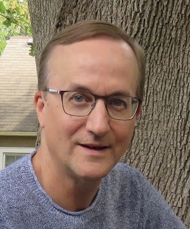 Photo of John Geddes in front of a tree trunk. John is wearing a grey shirt and glasses. His hair is short and brown. John is looking at the camera with a smile.