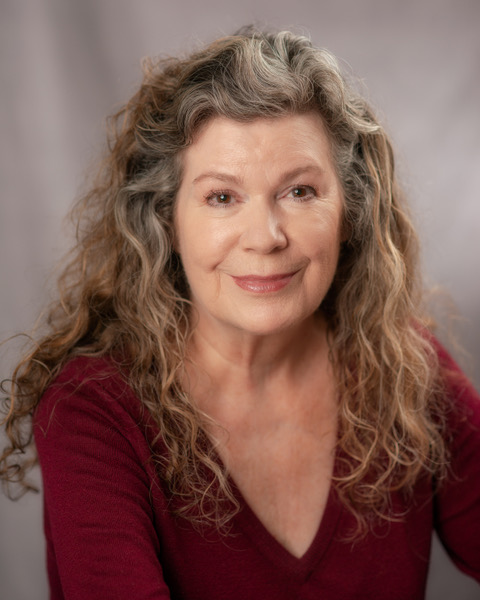 Photo of Laurie Koensgen in front of a light grey background. Laurie is wearing a red v-neck sweater, and her hair is long and curly. Laurie is looking at the camera with a smile.