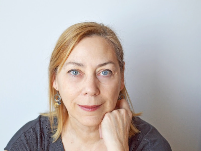 Photo of Susan Gillis in front of a white background. Susan is wearing a blue-grey shirt, and resting her chin on her closed hand. Susan is looking at the camera with a smile.