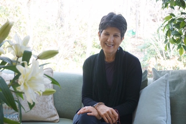 Photo of Jane Schapiro sitting on a grey couch with many pillows. Jane has her hands crossed with one hand resting on her knee. In the background is an overexposed forest, and in the foreground is a white flower.