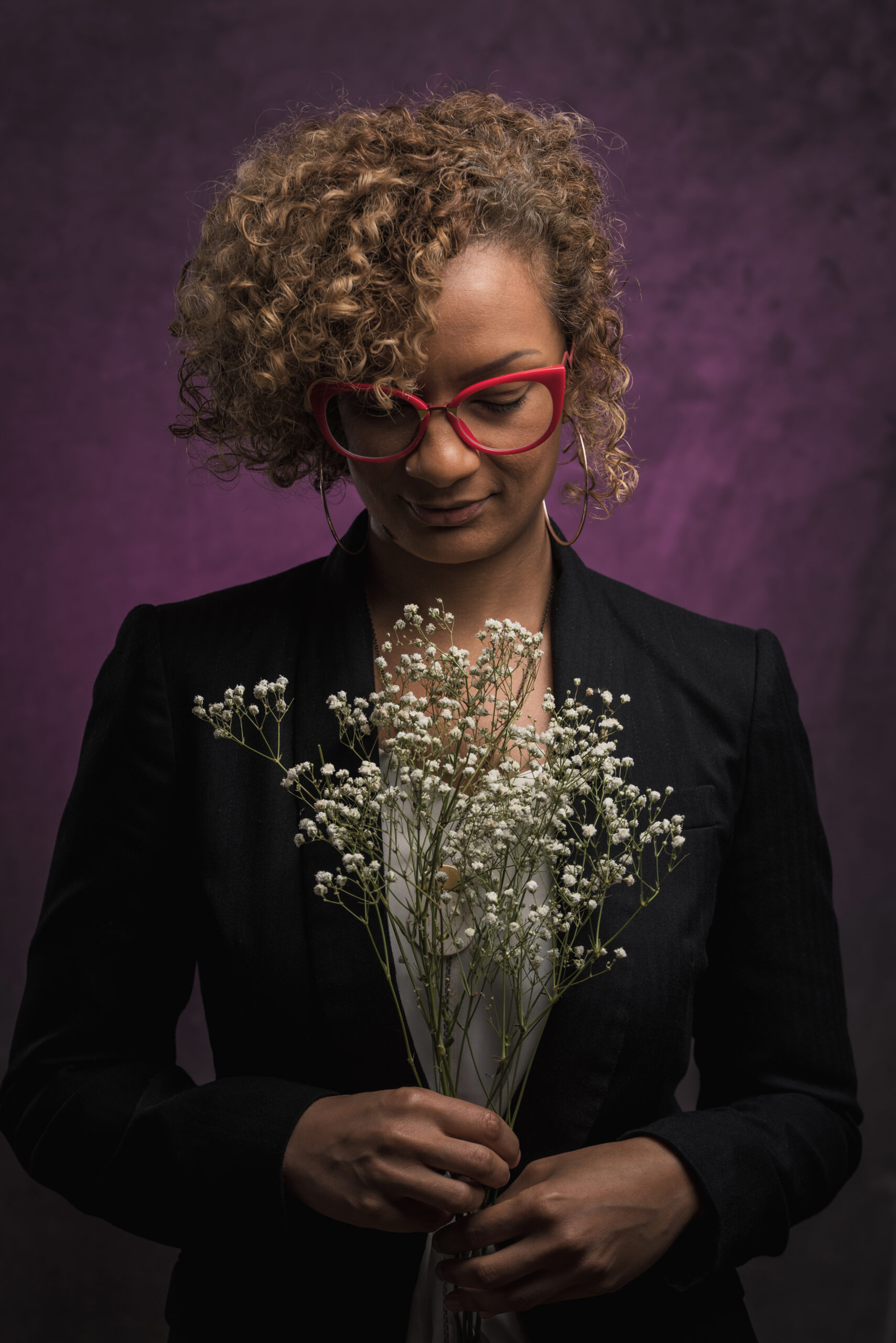Photo of Chelene Knight against a purple background. Chelene is wearing red glasses and a black blazer, and she is looking downward at a bouquet of flowers.