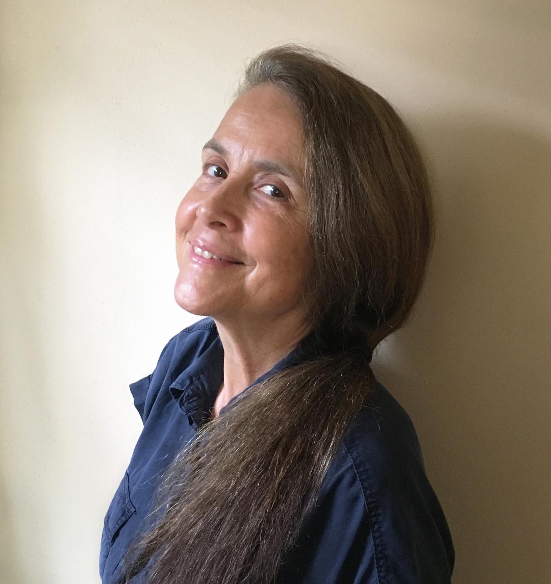 Photo of Naomi Shihab Nye leaning against a light tan wall. Nye is wearing a blue collar shirt and is smiling at the camera.