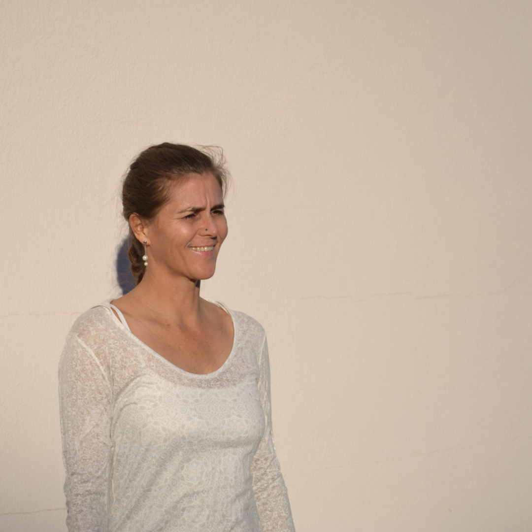 Photo of a woman in front of a pale beige or off-white wall. The woman is slightly off-centre, smiling and looking off to the right. Her long hair is pulled back in a ponytail.
